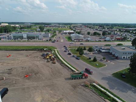 Aerial view of a construction site near a road intersection. Cars are visible on the roads beside the site. Nearby, various buildings and greenery surround the area under a partly cloudy sky.