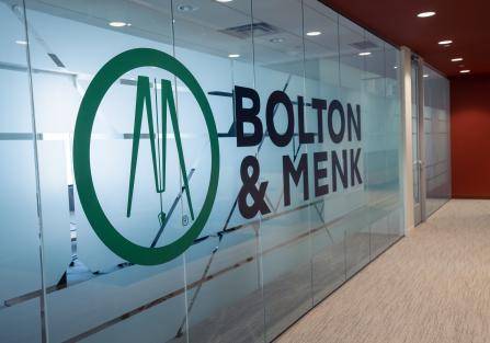 Glass office wall with BOLTON & MENK logo in black and a green circle emblem. The hallway is carpeted with beige flooring, and the left area shows a glimpse of a modern office interior.