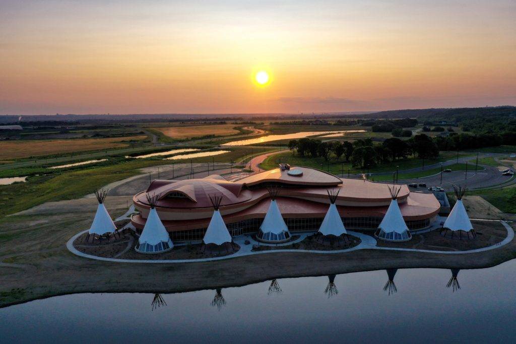 Aerial view of a building with a curved roof surrounded by tipi structures exemplifies innovative landscape architecture. Nestled beside a reflective body of water, the scene captures grasslands and a winding road as the sun sets, casting a warm glow over this harmonious design.