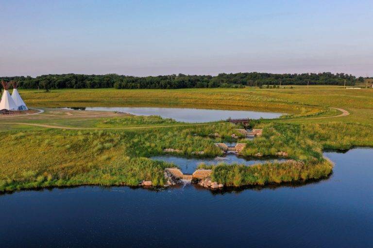 Aerial view of a tranquil landscape showcasing landscape architecture, with a teepee near a calm pond and rocky waterfalls. The area is lush and green under the blue sky, framed by a line of trees in the distance.