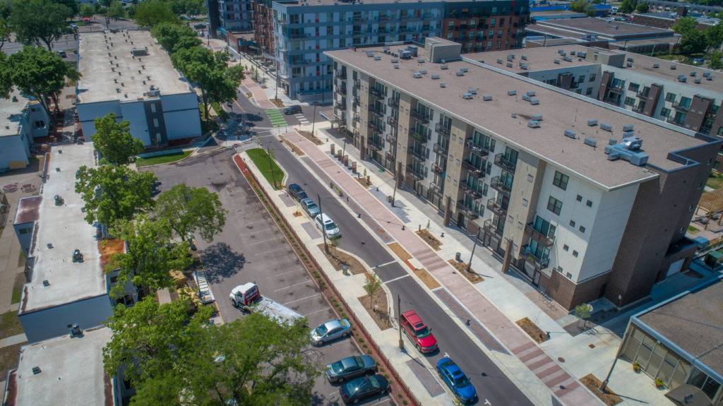An aerial view reveals a residential area with multi-story apartment buildings, showcasing elements of landscape architecture. Balconies and flat roofs overlook a tree-lined street, while parked cars dot the small parking lot surrounded by verdant trees.