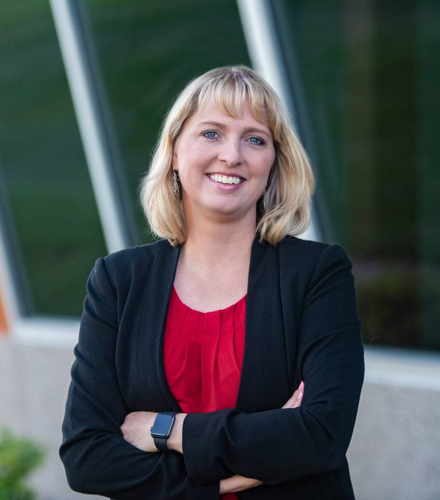 A woman with blonde hair in a black blazer and red blouse, representing the MN Young American Leaders Program, smiles confidently with arms crossed. She stands poised in front of a building with large windows.