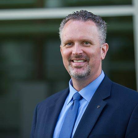 A person with short curly hair and a beard smiles while wearing a blue suit and tie, embodying the professional spirit of the NSPE Board of Directors. They stand confidently in front of a blurred background, possibly a building facade.