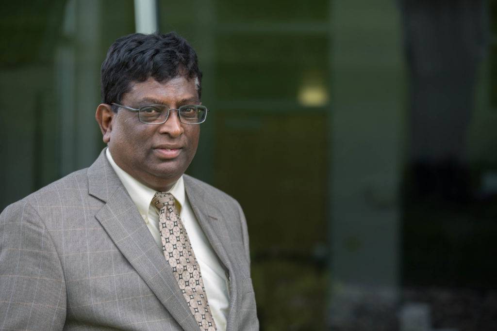 Herman Dharmarajah, in a plaid blazer and patterned tie, stands confidently in front of a glass building. With his glasses and short hair, he exudes an air of accomplishment, having recently received AWWA recognition. The blurred background highlights his poised presence.