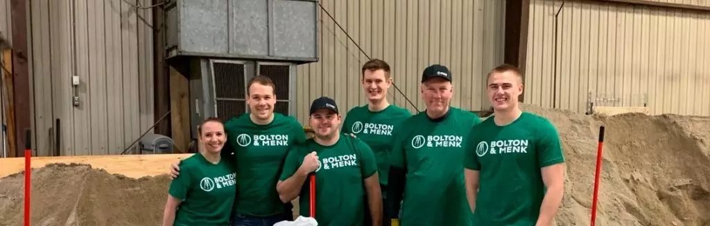 A group of six people wearing matching green T-shirts with Bolton & Menk stand indoors in front of a pile of gravel or sand, smiling and posing with shovels. Their enthusiasm highlights why Bolton & Menk is one of the Top 50 Best Places to Work.