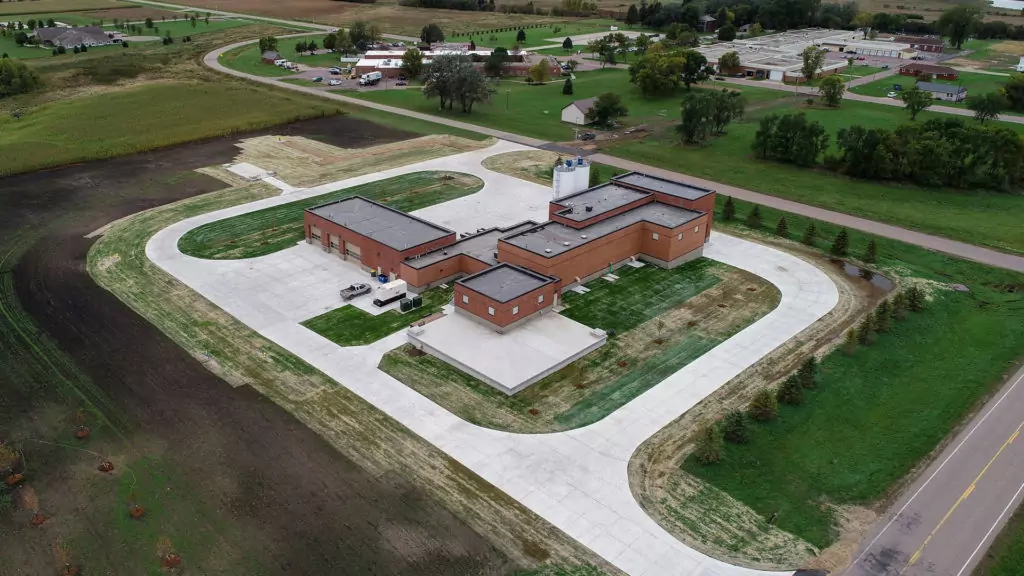 Aerial view of a large, U-shaped brick building with multiple driveways and parking areas, surrounded by grassy fields where efforts are made in reducing chlorides in wastewater. Nearby, various roads and buildings punctuate the rural landscape of trees and farmland.