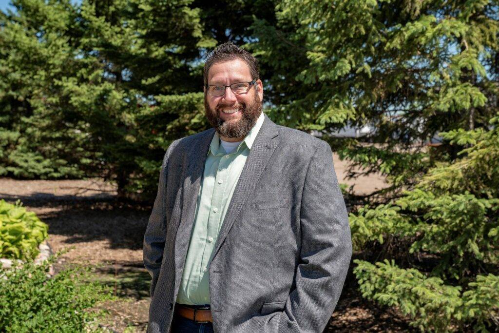 Chris Dahl, selected for Prairie Business 40 Under 40, beams with pride as he stands outdoors among pine trees. His beard and glasses add character to his look, complemented by a gray blazer over a light green shirt. The scene is set against lush greenery and a clear blue sky.