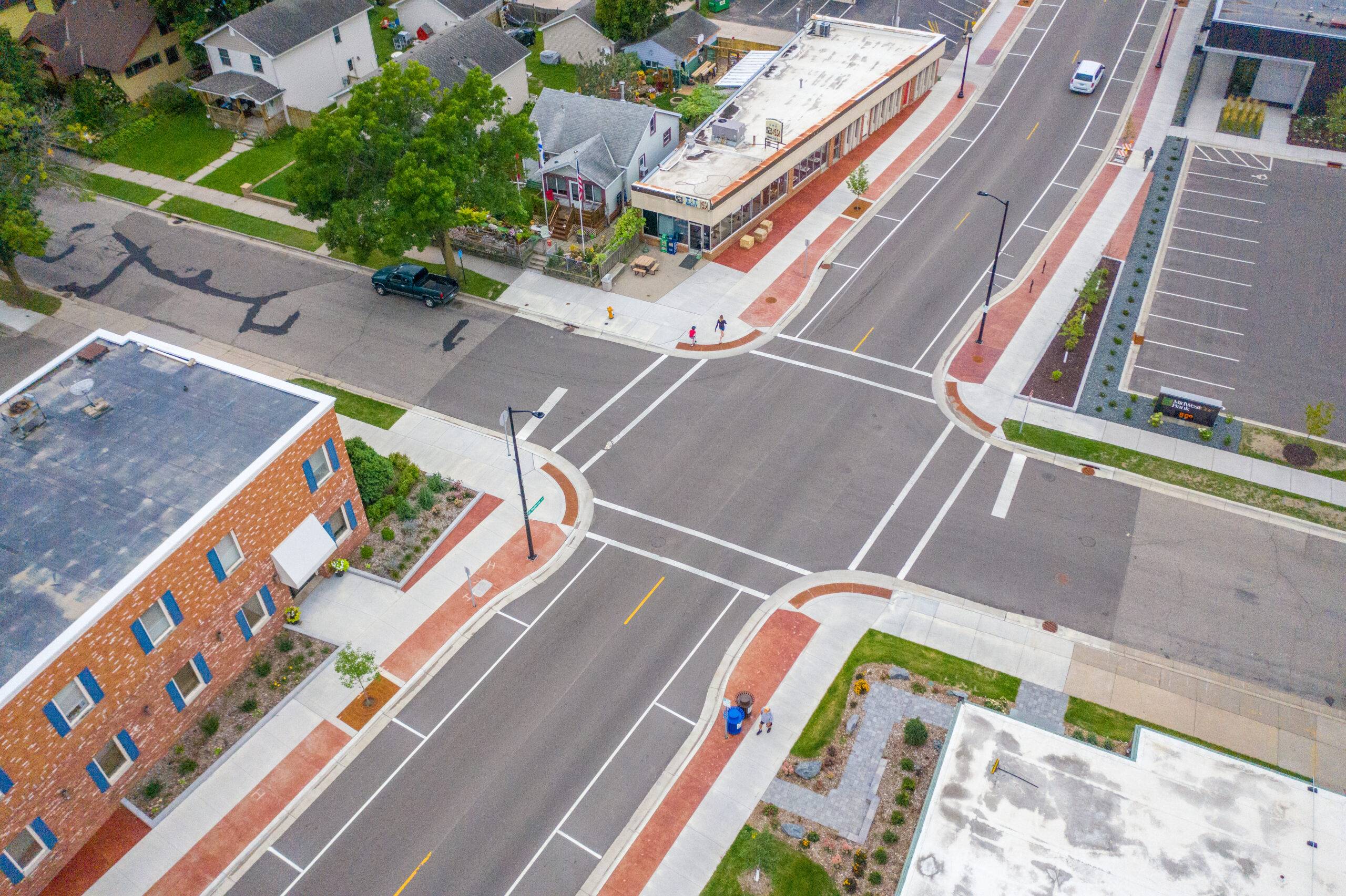 Aerial view of a city intersection with marked crosswalks and sidewalks, showcasing ADA-compliant solutions. Surrounding buildings include residential homes, a commercial structure, and parking areas. Streets are lined with trees and green spaces, with two people walking at the corner.