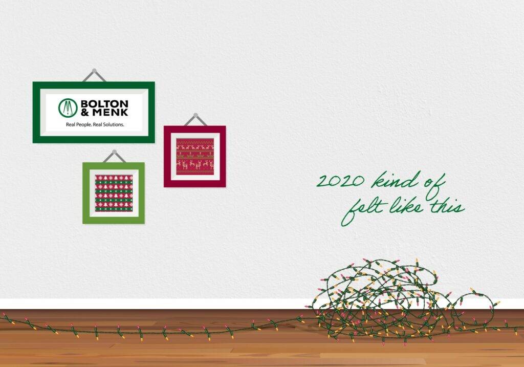 Two picture frames hang on a white wall, one with the Bolton & Menk logo and another showcasing holiday patterns. A tangled string of Christmas lights sprawls on the floor. The text whimsically reads, 2020 kind of felt like this. Happy Holidays!.