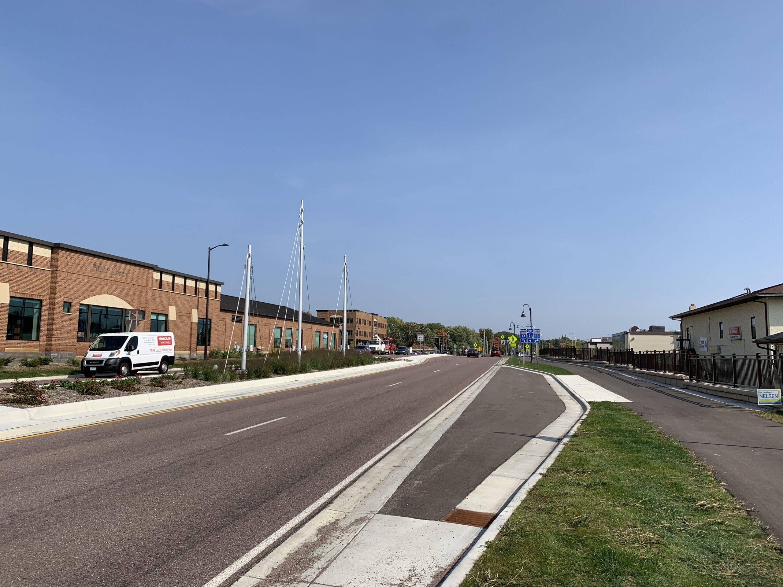 A wide, empty road in a suburban area with bike lanes on both sides suggests the thoughtful planning of the City of Prior Lake Downtown Project, which earned CEAM Project of the Year Honorable Mention. Modern brick buildings and greenery line the street under a clear blue sky.