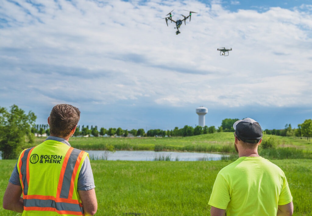 Two men stand in a grassy field, one wearing a safety vest labeled Bolton & Menk, as they operate two drones flying in the sky over a pond with a water tower in the distance on a partly cloudy day.
