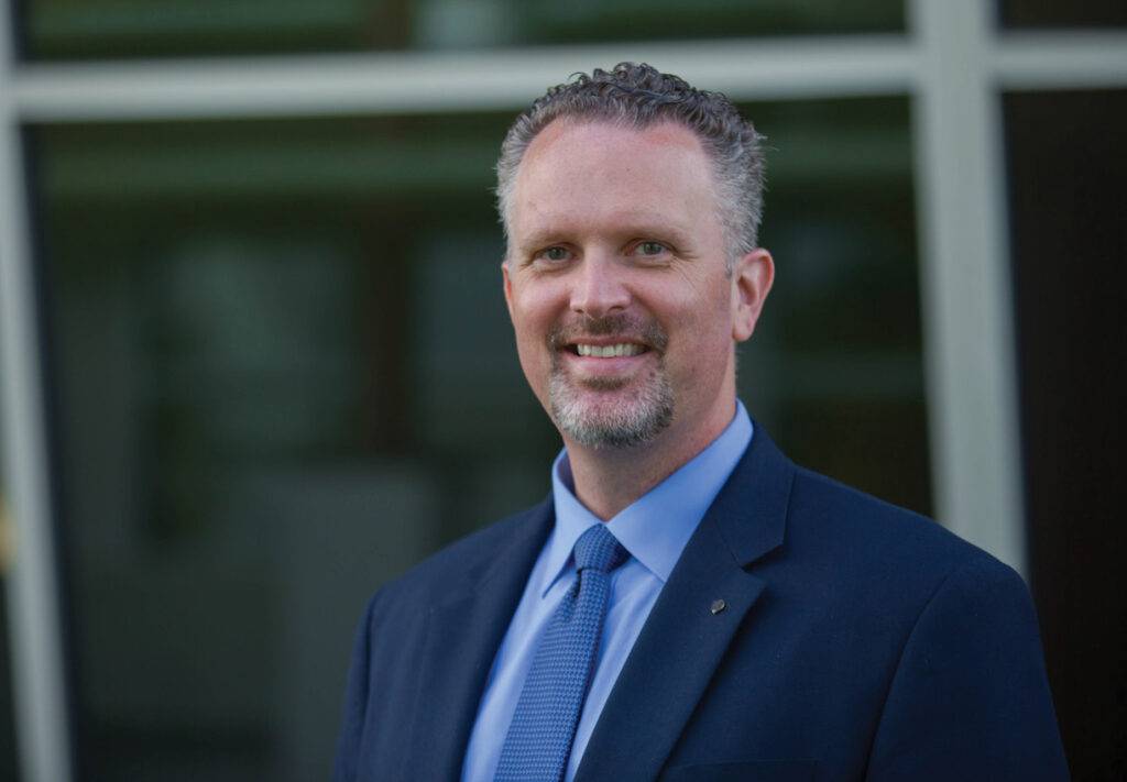 A smiling man with short, curly hair and a beard, Malm Named NSPE Fellow, stands confidently in front of a building with large windows, wearing a suit jacket, blue shirt, and tie.