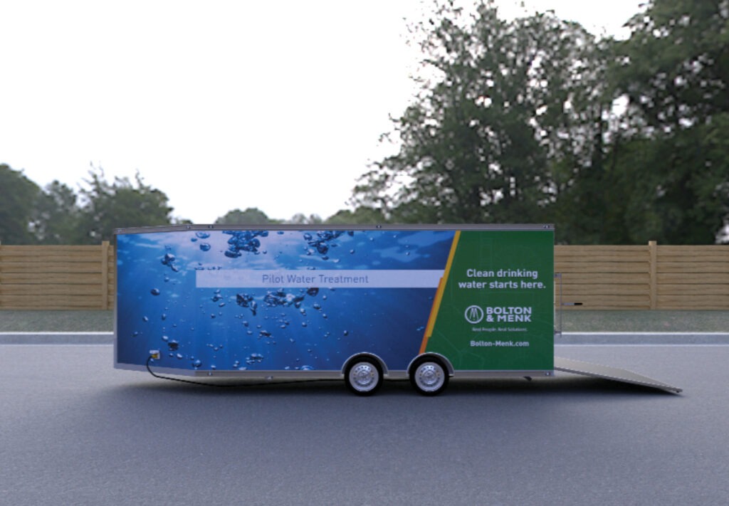 A trailer labeled “Pilot Water Treatment Plant” with water droplets and the Bolton & Menk logo is parked on a road beside a wooden fence and trees. The ramp is open. The slogan reads, Clean drinking water starts here.