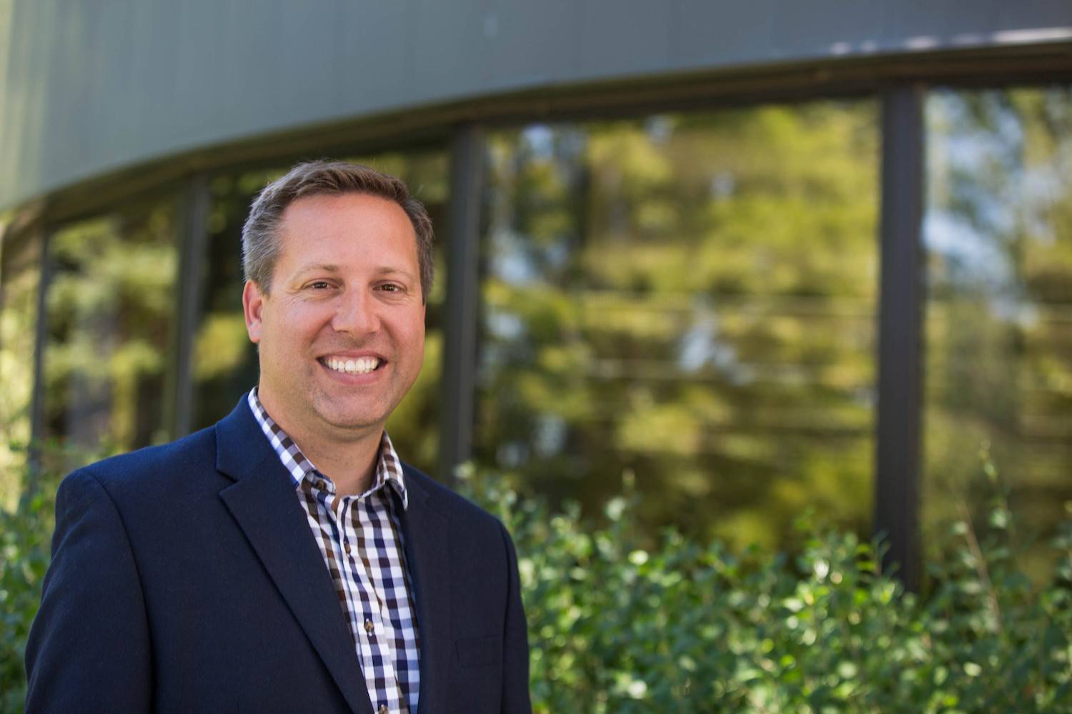 A person in a dark blazer and checkered shirt smiles while standing outdoors in the City of Anoka. Green foliage and a building with large windows are visible in the background.