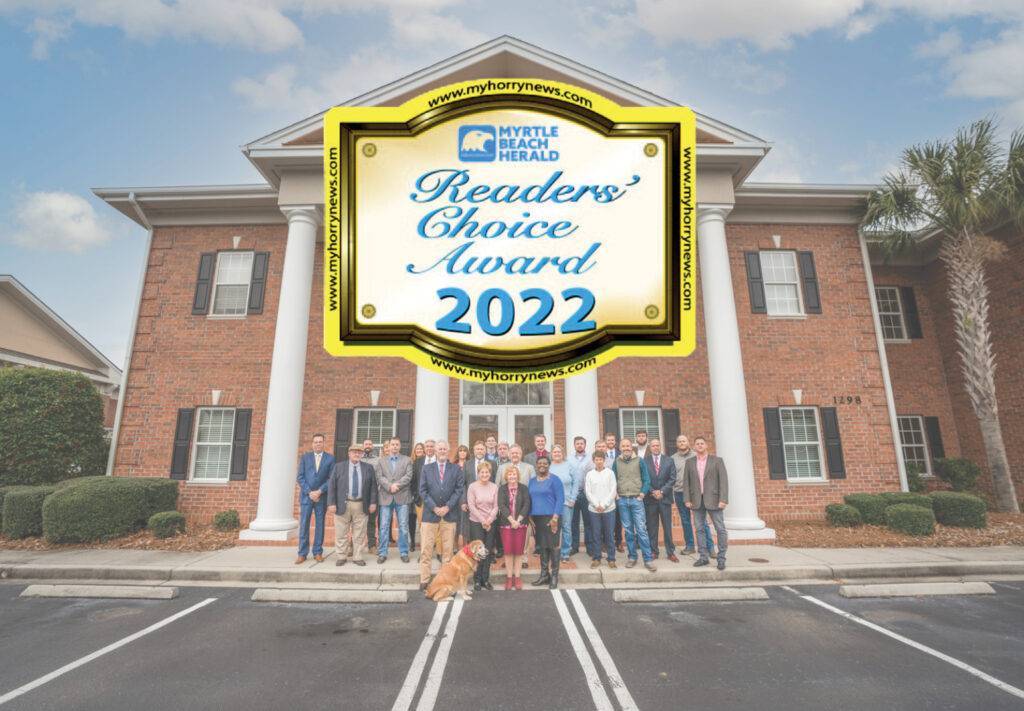 A group of people and a dog stand proudly in front of a brick building adorned with white columns. Overhead, a large Readers Choice Award 2022 badge from Myrtle Beach Herald graces the scene, celebrating their accomplishment.