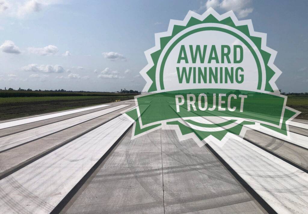 Concrete road stretch near Jefferson Municipal Airport, with a large green badge overlay that reads Award Winning Project set against a clear sky and lush green fields.