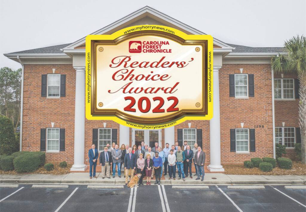 A group of people stands before a brick building with white columns, posing beneath a gold and red Carolina Forest Chronicle Readers Choice Award 2022 sign. A cheerful dog sits in front, embodying the spirit of the Carolina Forest Chronicle community.