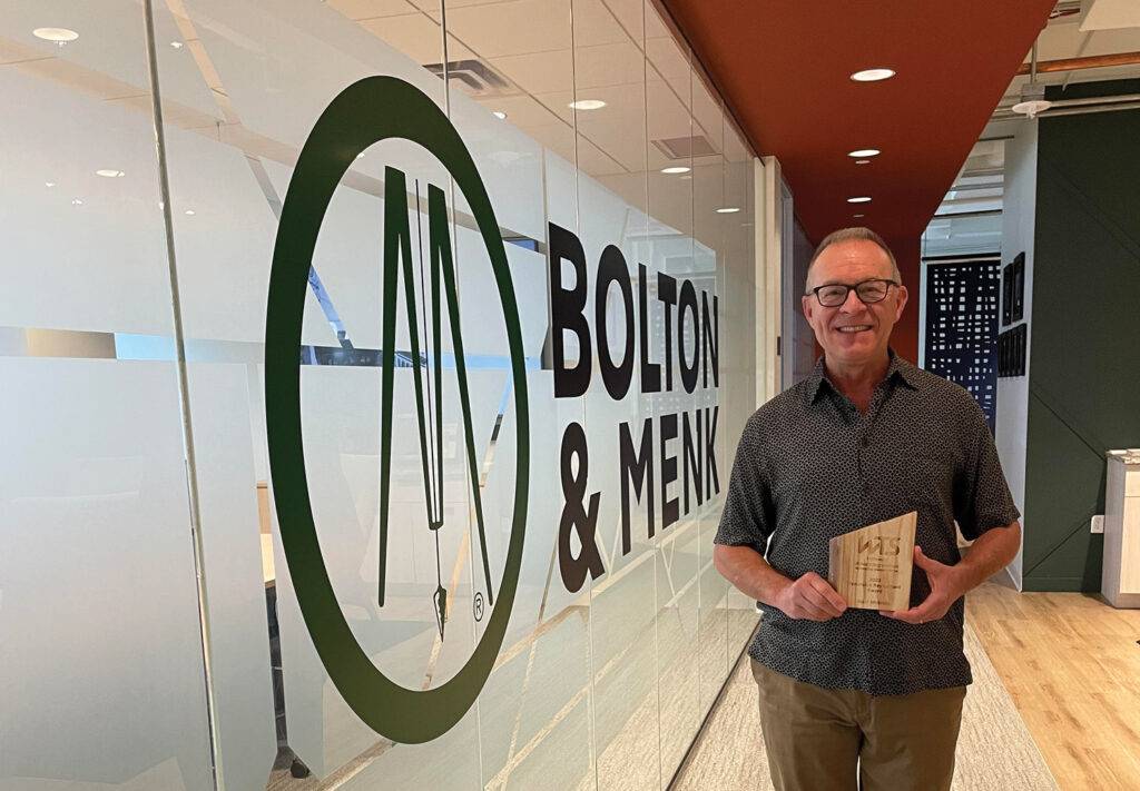 A person stands smiling in an office corridor next to a glass wall with the Bolton & Menk logo, proudly holding a wooden plaque for the Honorable Ray LaHood Award. The corridor boasts a modern design with light wood flooring and a striking red-accented wall.
