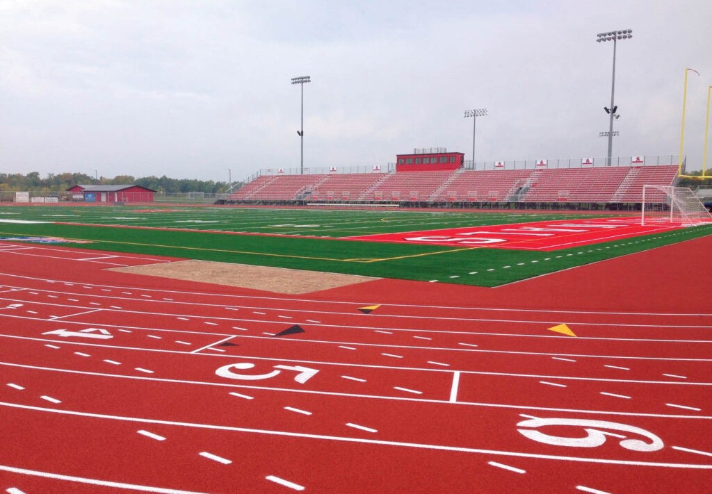 A red running track surrounds a green football field with red end zones, designed by Performance Sports Fields. Bleachers and stadium lights are visible in the background under a cloudy sky. The field and track are empty.