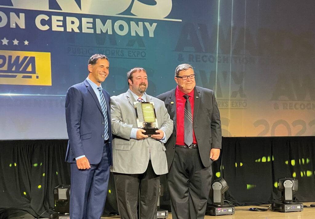 Three men in suits stand on a stage during the Professional Manager of the Year awards ceremony. The man in the middle proudly holds a trophy as the backdrop displays text related to the event.