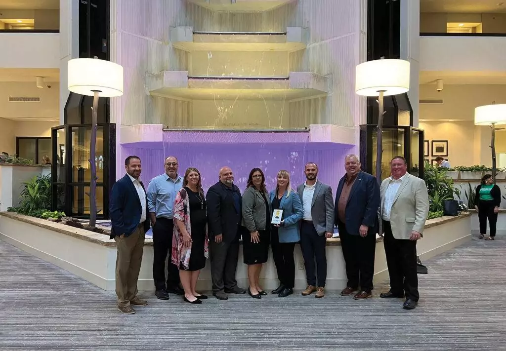 A group of ten people stands in front of a large, illuminated waterfall feature inside a building. They are dressed in formal attire and are smiling at the camera. The setting includes plants and modern lighting fixtures.