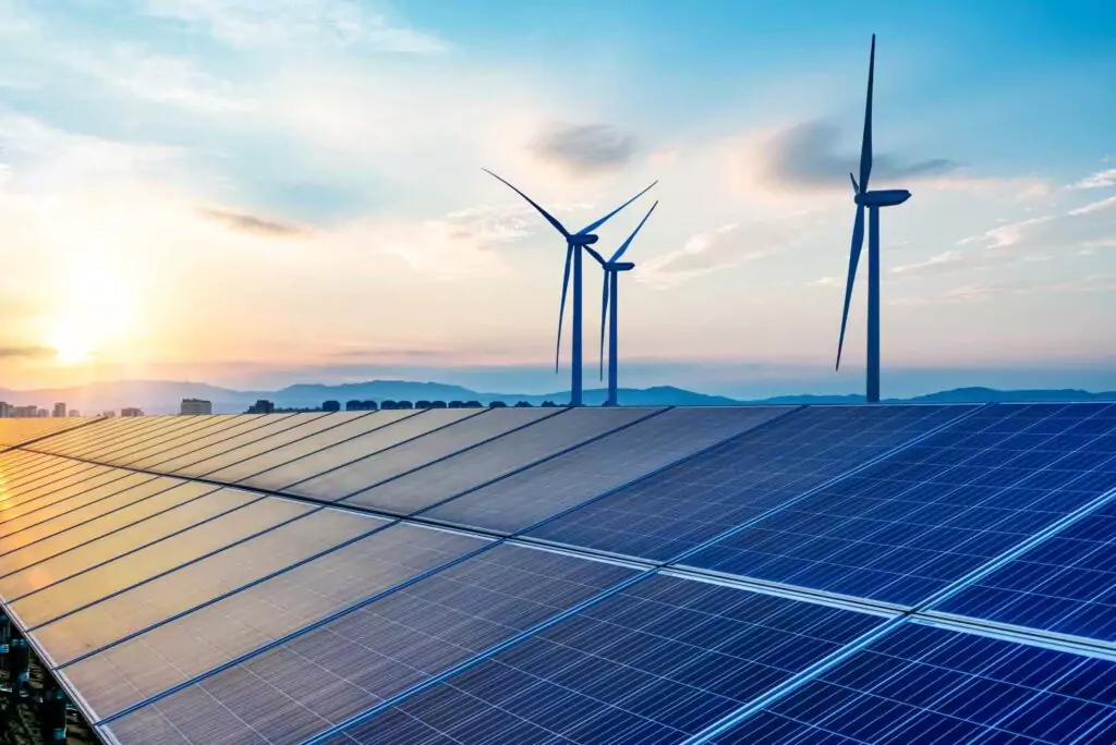 Solar panels and wind turbines are set against a vibrant sunrise, symbolizing renewable energy. The panels dominate the foreground, with the turbines and a cityscape visible in the background beneath a colorful sky.