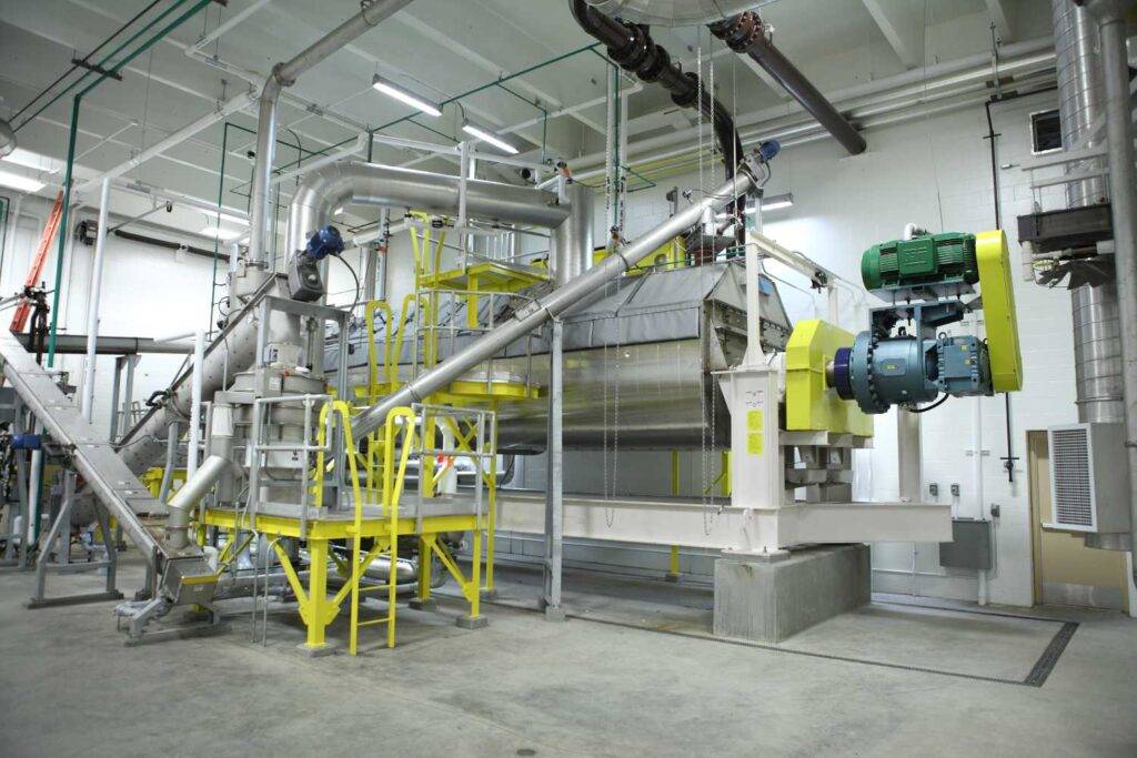 Industrial machinery set up in a factory with metal pipes, conveyor belts, and a large motor. The interior has white walls, overhead lighting, and safety railings painted yellow. The equipment is positioned on a concrete floor.