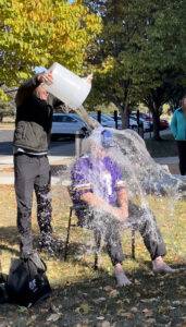 A person wearing a black shirt and hat pours a large bucket of water over another individual seated in a chair, donning a purple sports jersey. This playful volunteering event causes water to splash everywhere, amidst the backdrop of trees and a parked car.