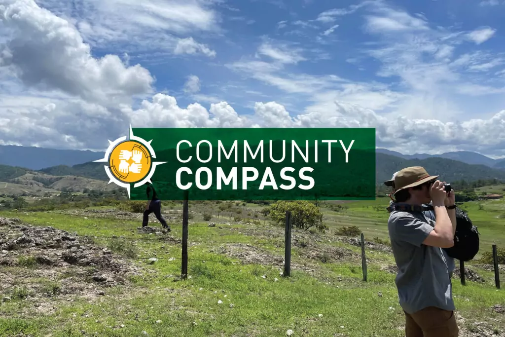 Two people are observing a grassy field under a blue sky with scattered clouds. One person uses binoculars, embodying the spirit of exploration akin to Engineers Without Borders. The scene is overlaid with a green banner displaying Community Compass and an icon of hands holding a compass.