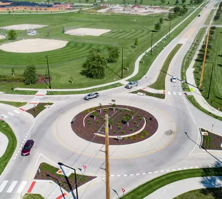 Aerial view of a traffic roundabout surrounded by green grass and landscaped areas. Nearby, a baseball field and other open spaces are visible. Cars are navigating the roundabout, and streets lead in multiple directions.