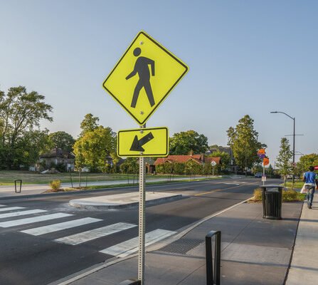 A pedestrian crosswalk sign on a two-lane road with a sidewalk in the background.