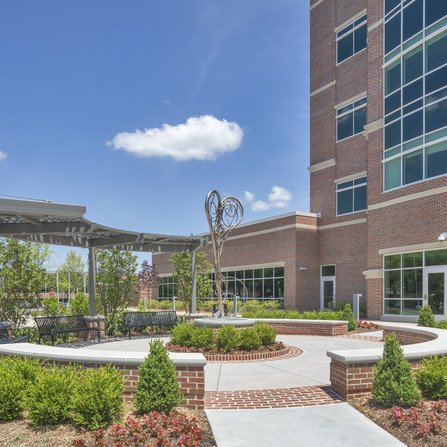 A modern brick office building with large windows under a clear blue sky. In front, there is a circular seating area with a contemporary sculpture. Green bushes and red flowers line the walkway leading to the entrance.