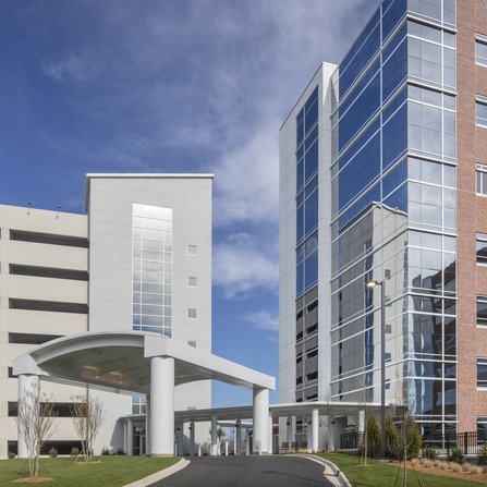 Modern hospital entrance with tall glass buildings on either side and a curved canopy covering the driveway. The sky is clear with a few clouds, and there are patches of grass near the path.