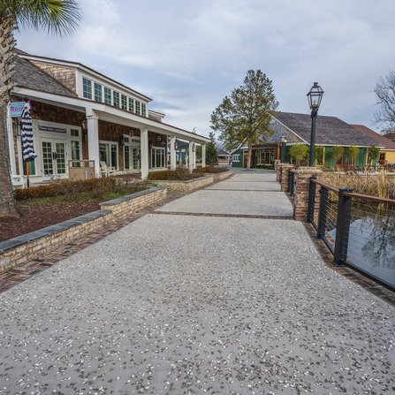 A quiet street lined with rustic buildings features a tree and a black lamppost on the right. The sky is overcast, and a palm tree stands near the left building. A stone pathway leads between the structures, bordered by a low wall on the left.