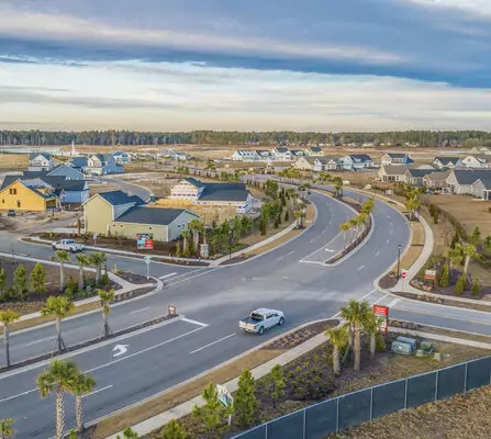 Aerial view of a suburban residential area with a curving road, lined with neatly arranged houses and manicured gardens. A single car travels on the road. The sky is partly cloudy, and there is a fenced-off area in the foreground.