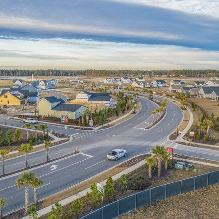 Aerial view of a suburban residential area with a curving road, lined with neatly arranged houses and manicured gardens. A single car travels on the road. The sky is partly cloudy, and there is a fenced-off area in the foreground.