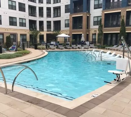 A modern apartment complex surrounds a rectangular outdoor pool with clear blue water. Lounge chairs and umbrellas line the poolside, and greenery decorates the area. The building features a mix of white and brick facade.