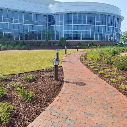 Curved brick pathway leading to a modern building with large glass windows. The path is lined with small black lamp posts, surrounded by neatly trimmed grass and young shrubs. The building has a cylindrical structure.