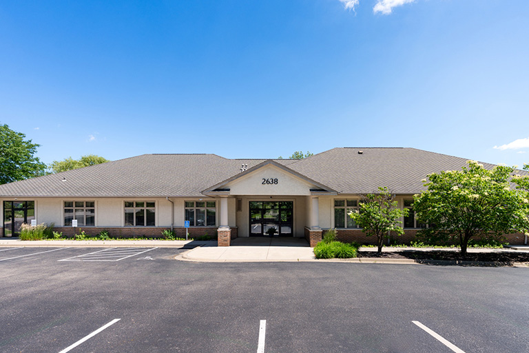 A single-story building with a gray roof, light-colored walls, and the number 2638 above the entrance. It is surrounded by trees and greenery, with a large empty parking lot in the foreground. The sky is clear and blue.