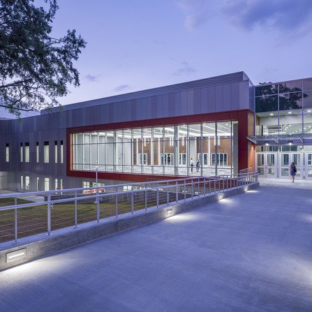 Modern building with large glass windows and a prominent red frame around a central section. A walkway with metal railings leads to the entrance. The scene is lit with soft evening lighting, and a tree is visible on the left.