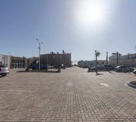 A sunlit, empty brick-paved parking lot with some parked cars on either side. A few palm trees are scattered around. In the background, a clear blue sky highlights the silhouette of a brick building.