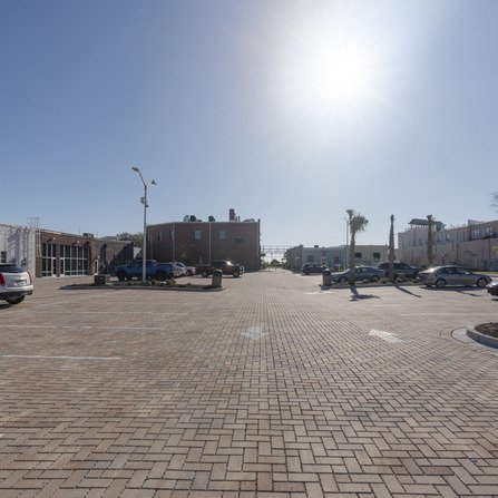 A sunlit, empty brick-paved parking lot with some parked cars on either side. A few palm trees are scattered around. In the background, a clear blue sky highlights the silhouette of a brick building.