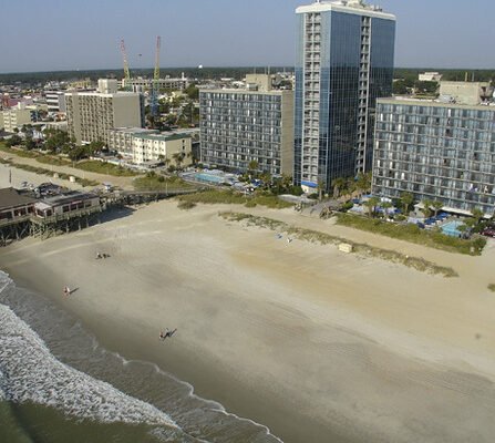 Aerial view of a coastal city with high-rise hotels along a sandy beach. The ocean waves gently crash onto the shore, and a few people are visible on the sand. Several cranes and city buildings are visible in the background.