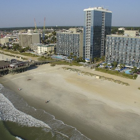 Aerial view of a coastal city with high-rise hotels along a sandy beach. The ocean waves gently crash onto the shore, and a few people are visible on the sand. Several cranes and city buildings are visible in the background.