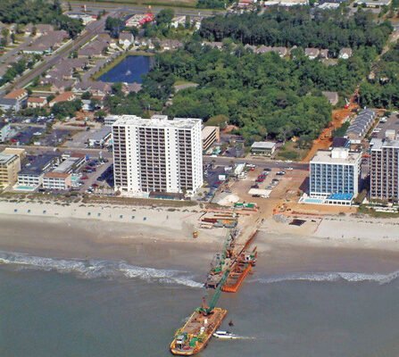 Aerial view of a coastal town with high-rise buildings along the shoreline. A dredging vessel operates near the beach, connected to the shore by a pipeline. The town has trees and residential areas visible inland.