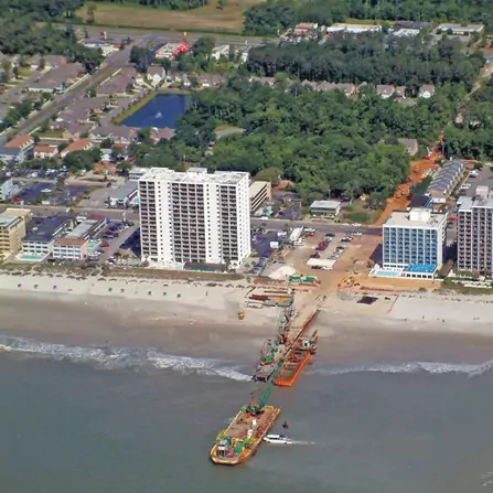 Aerial view of a coastal town with high-rise buildings along the shoreline. A dredging vessel operates near the beach, connected to the shore by a pipeline. The town has trees and residential areas visible inland.