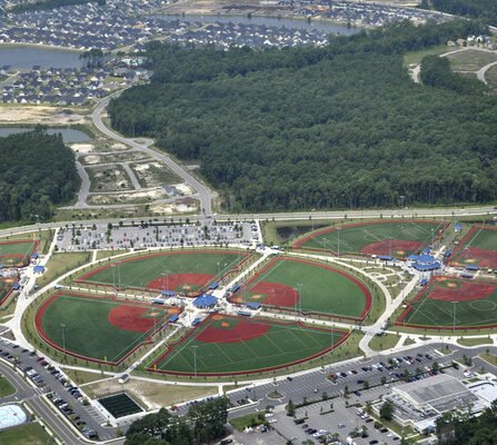 Aerial view of a sports complex featuring multiple circular fields, some with red and green surfaces, likely for baseball or softball. Surrounding areas include parking lots, buildings, and residential neighborhoods against a backdrop of trees.