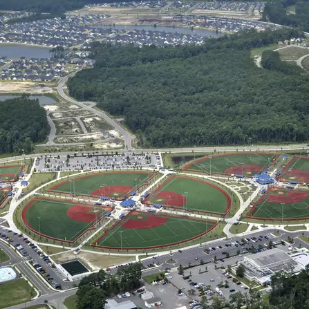 Aerial view of a sports complex featuring multiple circular fields, some with red and green surfaces, likely for baseball or softball. Surrounding areas include parking lots, buildings, and residential neighborhoods against a backdrop of trees.
