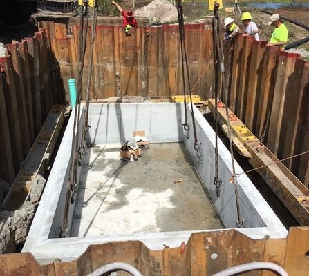 A large rectangular concrete pit in a construction site, secured with metal beams and surrounded by wooden barriers. Several workers in safety gear are visible in the background.