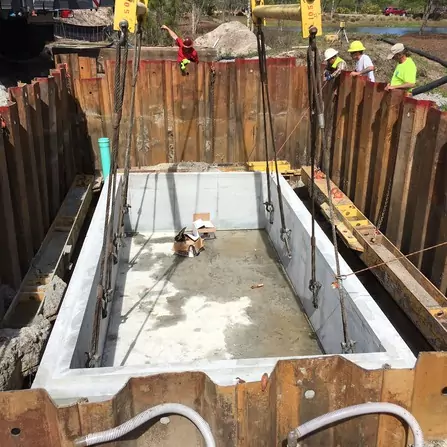 A large rectangular concrete pit in a construction site, secured with metal beams and surrounded by wooden barriers. Several workers in safety gear are visible in the background.